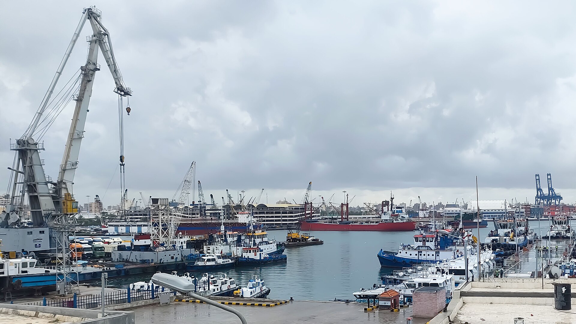 Cranes, tugboats and a cargo ship on the water at the Port of Alexandria, Egypt, under an overcast sky